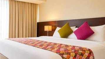 Habitación de hotel con una cama grande, cabecero, lámpara de noche y ventana con cortinas.