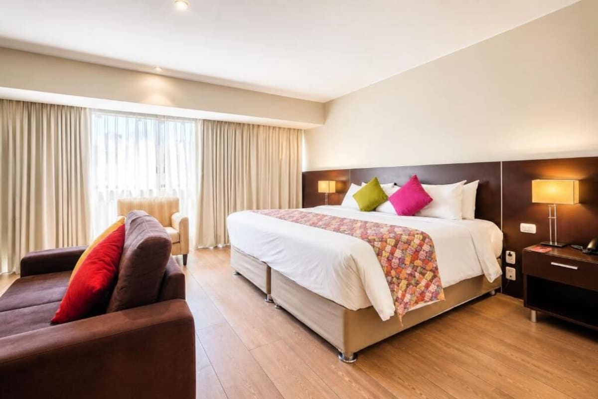 A hotel room with a large bed, a sofa, a nightstand, and a window with curtains.