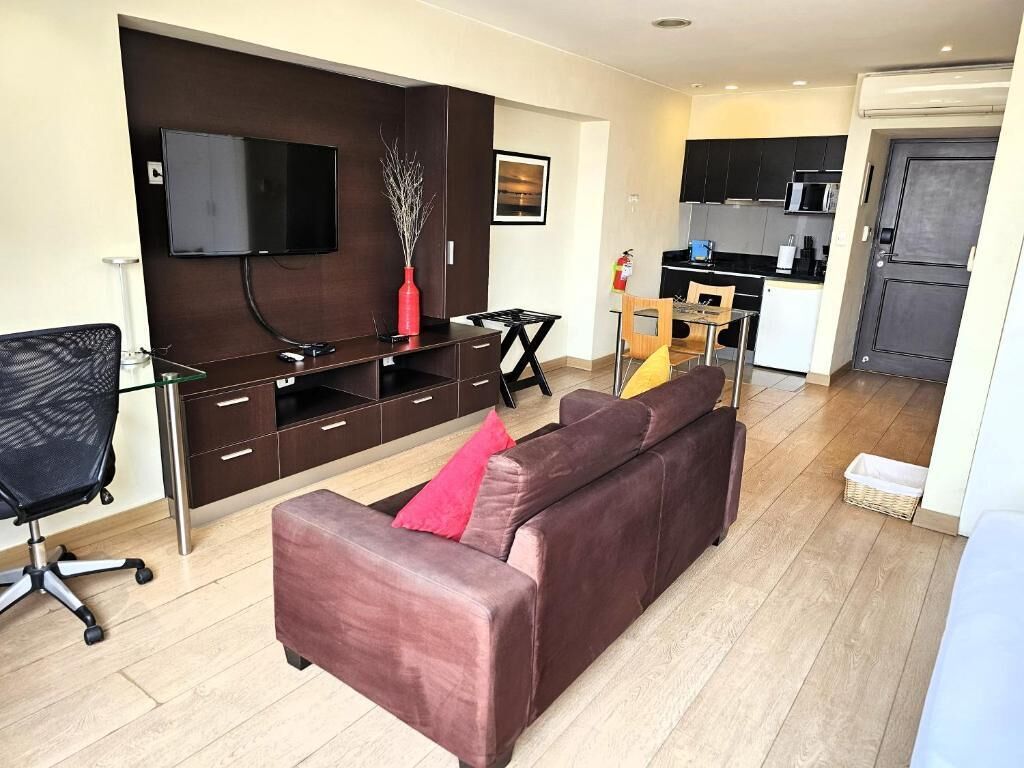 A modern living room with a brown sofa, a TV mounted on the wall, and a dining area with a table and chairs.
