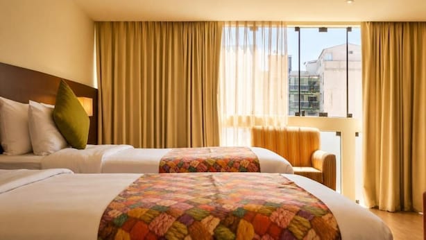 A hotel room with two beds, a sofa, and a view of buildings through large windows. - Diez Canseco 2W Apartments Service (Miraflores)