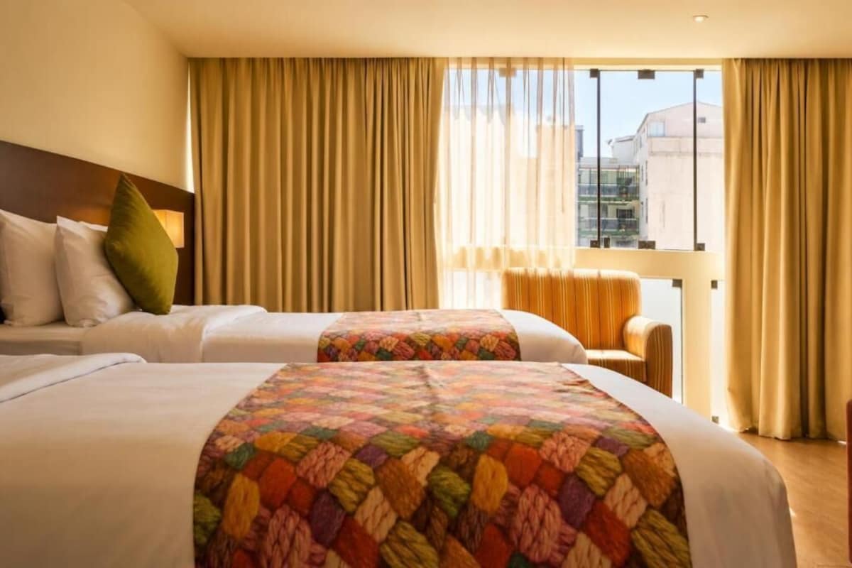 A hotel room with two beds, a sofa, and a view of buildings through large windows.