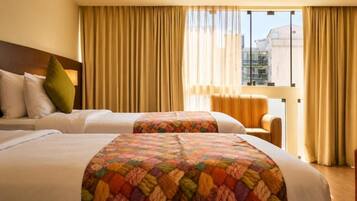 Habitación de hotel con dos camas, un sofá y vistas a edificios a través de amplios ventanales.