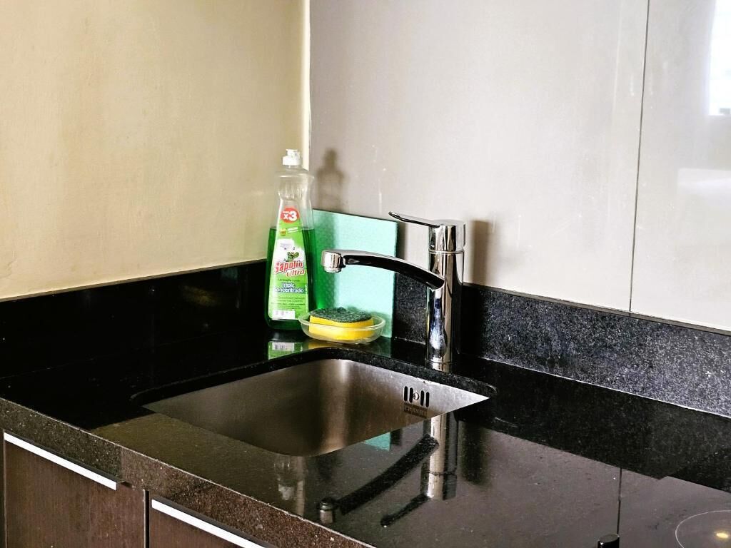 Una cocina con un lavamanos de acero inoxidable, una botella verde de lavavajillas y una esponja amarilla.