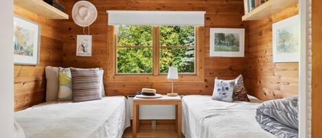 A cozy cabin room with two beds, a window with a view of greenery, and wooden walls.