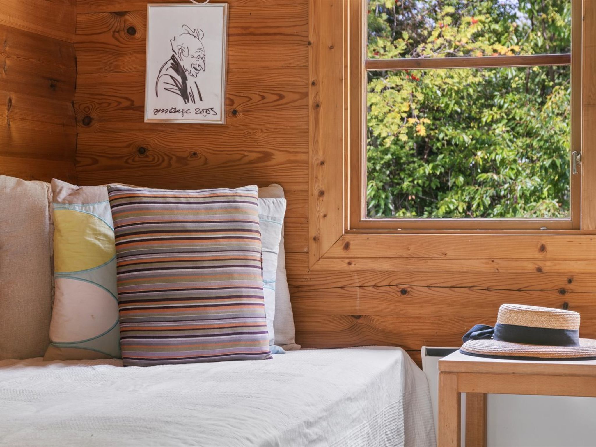 Interior de una cabaña de madera con una cama, almohadas, un dibujo enmarcado y una ventana con vista a follaje.