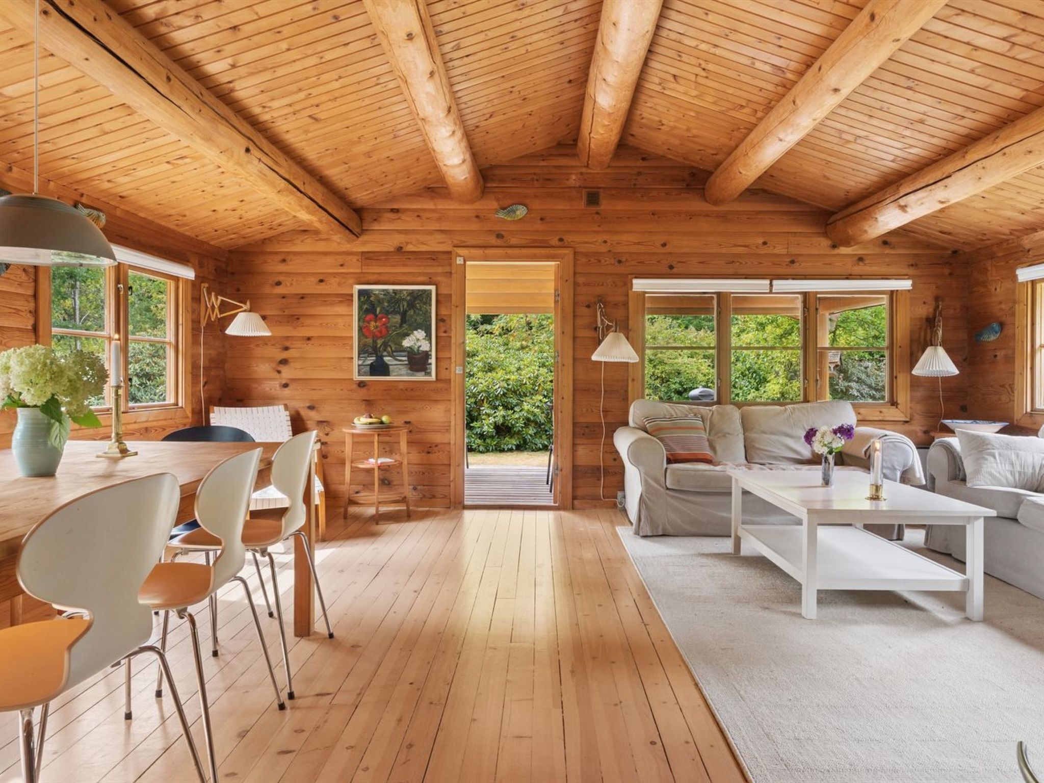 A wooden cabin interior with a dining area, a sofa set, and a view of greenery outside.