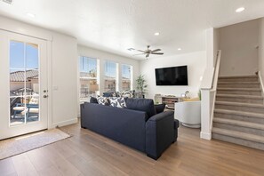 A living room with a dark sofa, a flat-screen TV, and a staircase. - Desert Oasis - Pool, Hot Tub and Pickleball Courts (Washington)