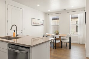 A modern kitchen with a central island, a dining area with a table and chairs, and a window with blinds. - Desert Oasis - Pool, Hot Tub and Pickleball Courts (Washington)