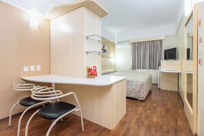Ein kompaktes Hotelzimmer mit Küchenzeile, einem Bett mit gemustertem Überwurf, einer Sitzecke mit Stühlen und einem Fernseher.