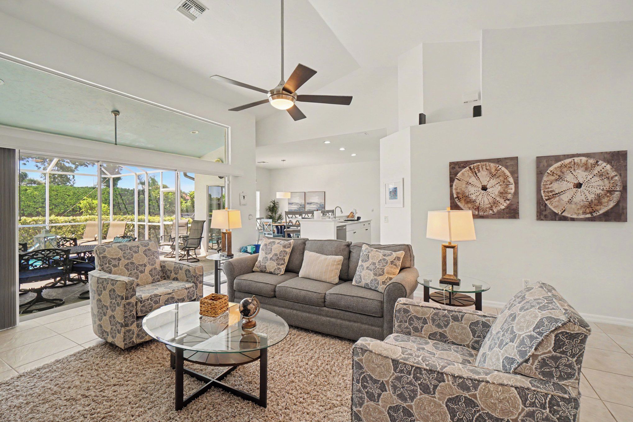 A spacious living room with a glass coffee table, a sofa, and armchairs, a ceiling fan, and a view of the outdoors.
