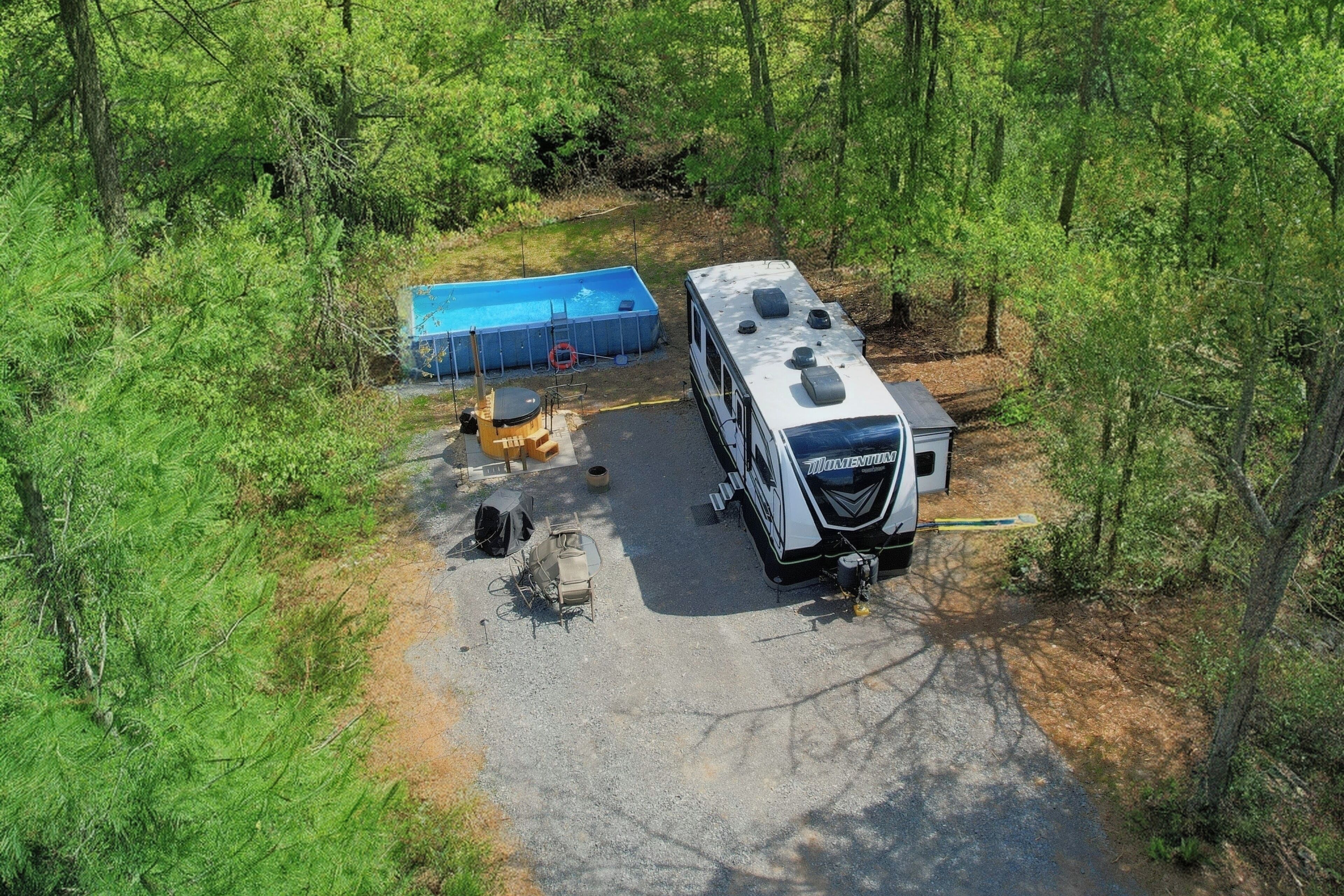 A recreational vehicle (RV) parked in a forested area with a swimming pool and outdoor seating.