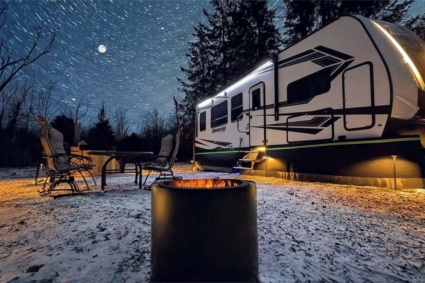 A camper RV parked outdoors at night with a fire pit in the foreground.