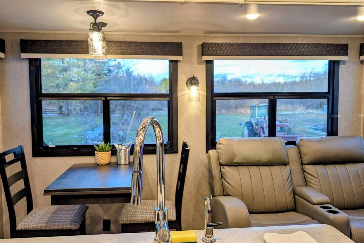 A cozy RV interior with a dining area, a sofa, and large windows offering views of a grassy area.