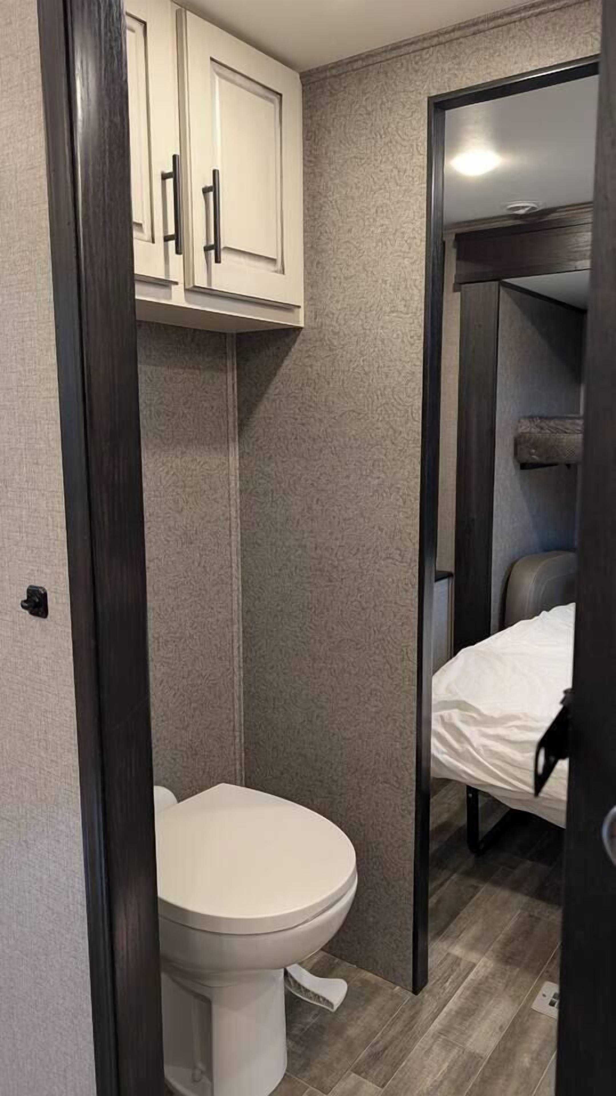 Une petite salle de bain comprenant des toilettes, un meuble de rangement au-dessus et un lit dans la pièce adjacente.
