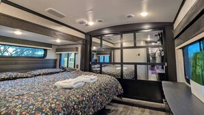 A modern RV bedroom with two beds, a large window, and a sliding door to the bathroom.