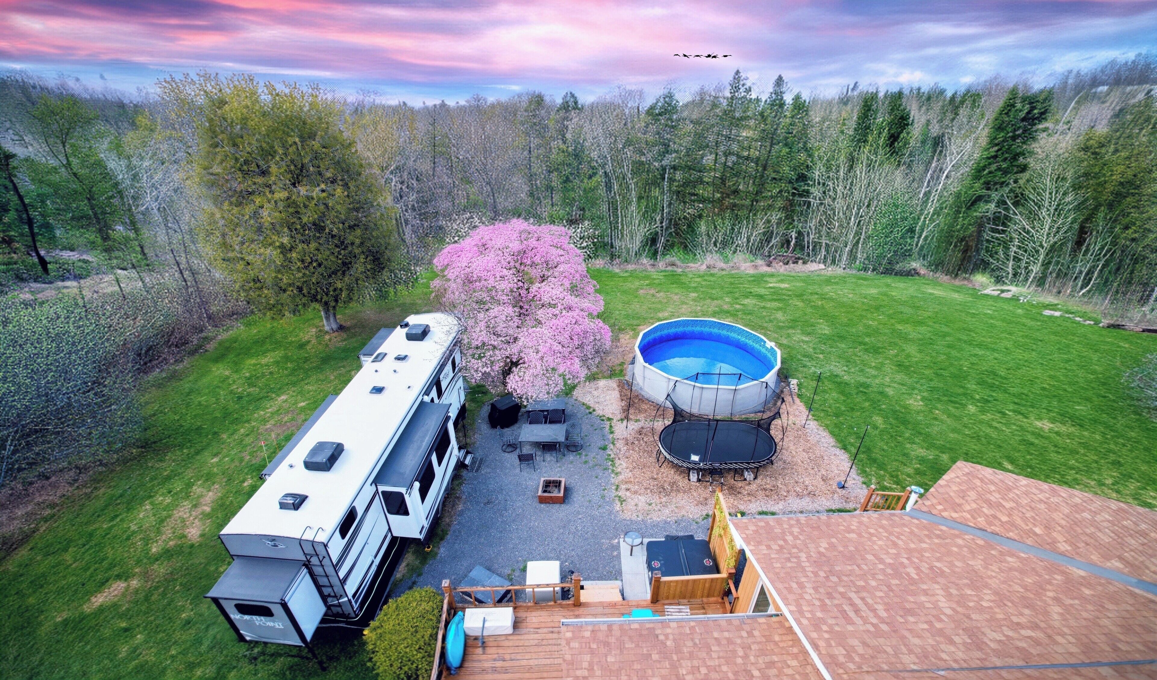 A recreational vehicle with a covered patio, a swimming pool, and a tree with pink blossoms.