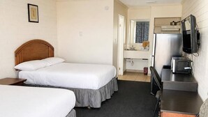 Quarto de hotel com duas camas de solteiro, uma pequena escrivaninha com cadeira, televisão e cozinha compacta.