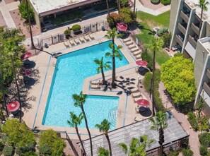 Eine Luftaufnahme eines Hotels mit einem rechteckigen Pool, umgeben von Liegestühlen und Sonnenschirmen, Palmen und einem Gebäude neben dem Poolbereich.