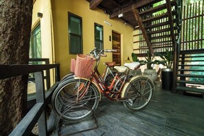 Un vélo est garé sur une terrasse en bois, avec un panier fixé à l’avant, près d’un bâtiment à la façade jaune et d’une clôture en bois.