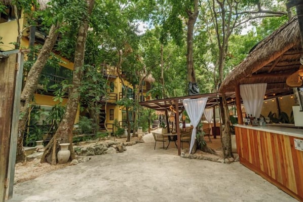 Un espace extérieur rustique doté d’un bar à toit de chaume, de sièges en pierre et d’une végétation luxuriante.