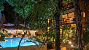 A pool area with lounge chairs and umbrellas, surrounded by tropical plants and a building with large windows.