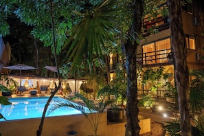 Un espace piscine agrémenté de chaises longues et de parasols, entouré de plantes tropicales et bordé d’un bâtiment aux grandes fenêtres.