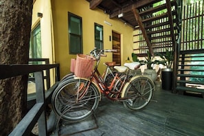 Un vélo est garé sur une terrasse en bois, avec un panier fixé à l’avant, près d’un bâtiment à la façade jaune et d’une clôture en bois.