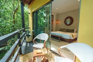 Een slaapkamer met een bed, nachtkastje en stoel. Er is een balkon met een hekwerk en een bloempot.