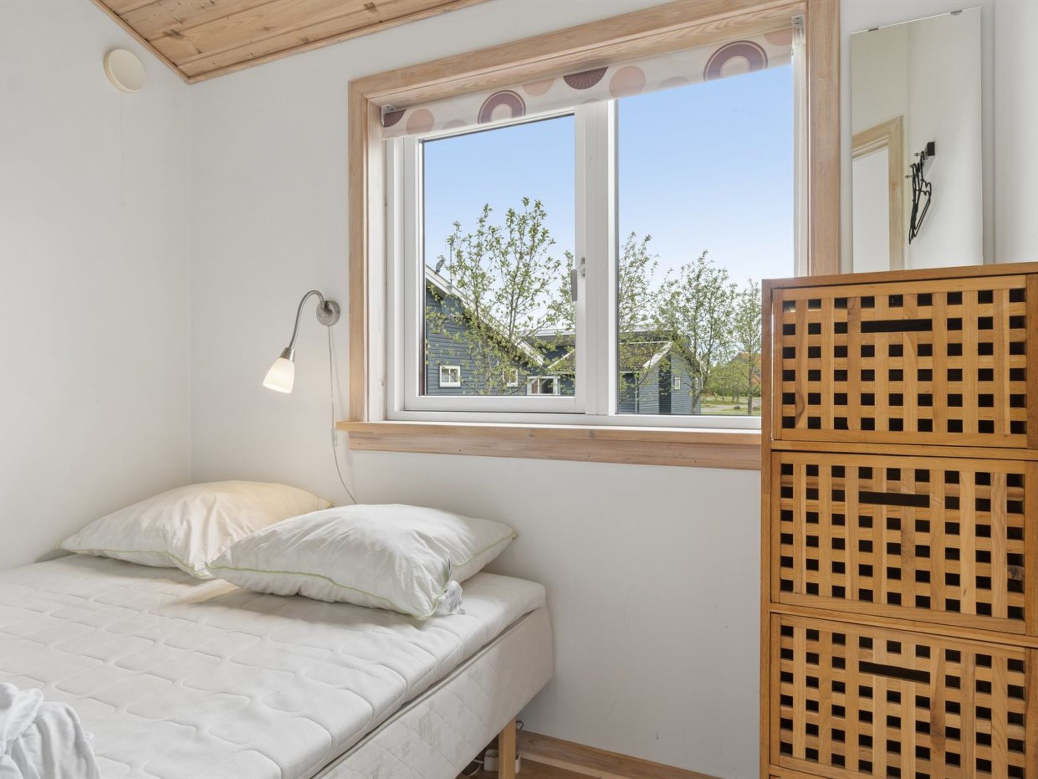 Une chambre avec un lit simple, une fenêtre donnant sur des arbres et une maison, et une armoire en bois.