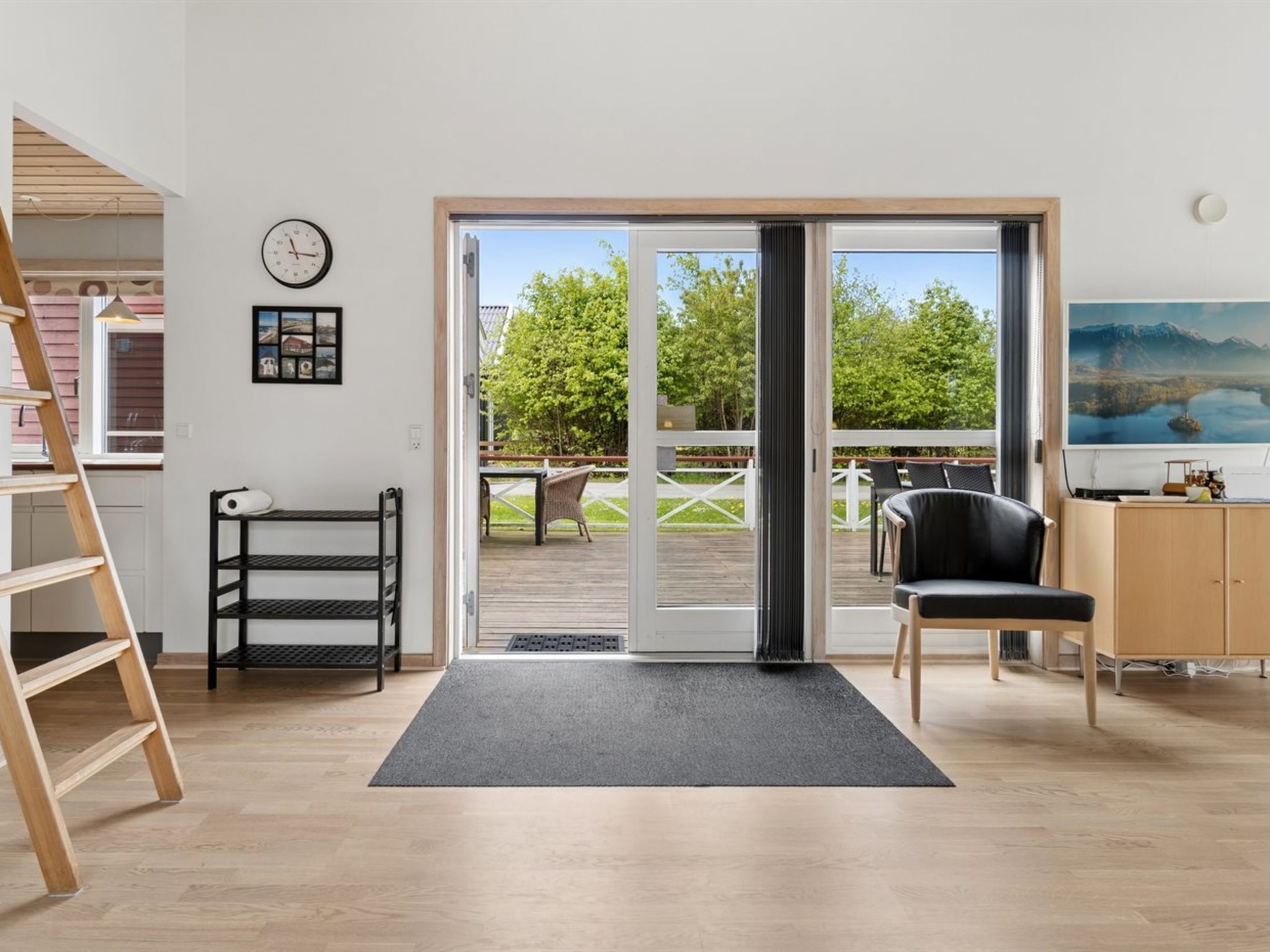Ein Zimmer mit Holzboden, einem Schreibtisch mit Stuhl, einem schwarzen Stuhl, einer schwarzen Regalwand, einer Uhr und einer Schiebetür zu einer Terrasse.