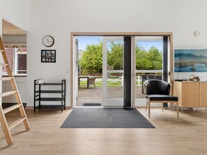A room with a wooden floor, a desk with a chair, a black chair, a black shelf unit, a clock, and a sliding glass door leading to an outdoor deck. - "Sannie" - 2.8km from the sea by Interhome (Gudhjem)