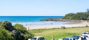 A beach with a grassy area and parked cars, a rocky outcrop in the water, and a clear sky. - Aqua - Front row living on Woolgoolga Beach (Woolgoolga)