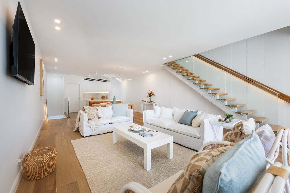 Una sala de estar moderna con una escalera, sofás blancos, una mesa de centro y un televisor de pantalla plana montado en la pared.