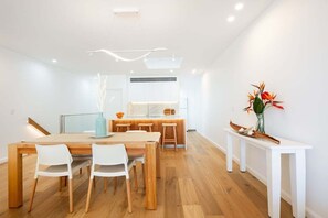 A modern dining area with a wooden table and white chairs, a kitchen in the background, and a white console table with a vase of flowers. - Aqua - Front row living on Woolgoolga Beach (Woolgoolga)