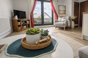 Een woonkamer met een televisie, een bank, een bijzettafeltje met planten en uitzicht op een houten terras door een glazen deur.
