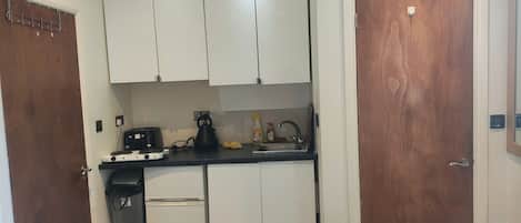 Una cocina compacta con armarios blancos, encimera negra, cafetera, hervidor y lavabo.