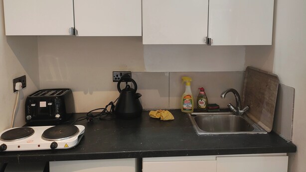 A kitchen with white cabinets, a black countertop, a kettle, a toaster, a sink, and cleaning supplies. - Studio Sleeps 2 with Parking (London)