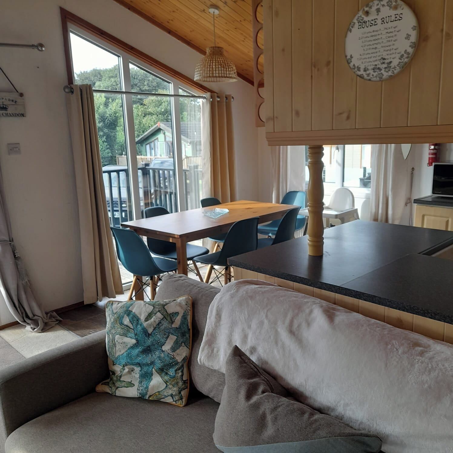 Ein gemütliches Zimmer mit Holzbalkendecke, einem Esstisch mit Stühlen, einer Couch mit Kissen und Blick ins Freie durch große Fenster.