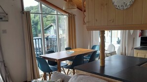 Une pièce chaleureuse avec un plafond en bois, une table à manger avec des chaises, un canapé avec des coussins et une vue sur l’extérieur grâce à de grandes fenêtres.