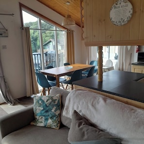 A cozy room with a wooden ceiling, a dining table with chairs, a sofa with cushions, and a view of the outdoors through large windows. - Cabin Sleeps 6 with Indoor Pool Parking (Looe)