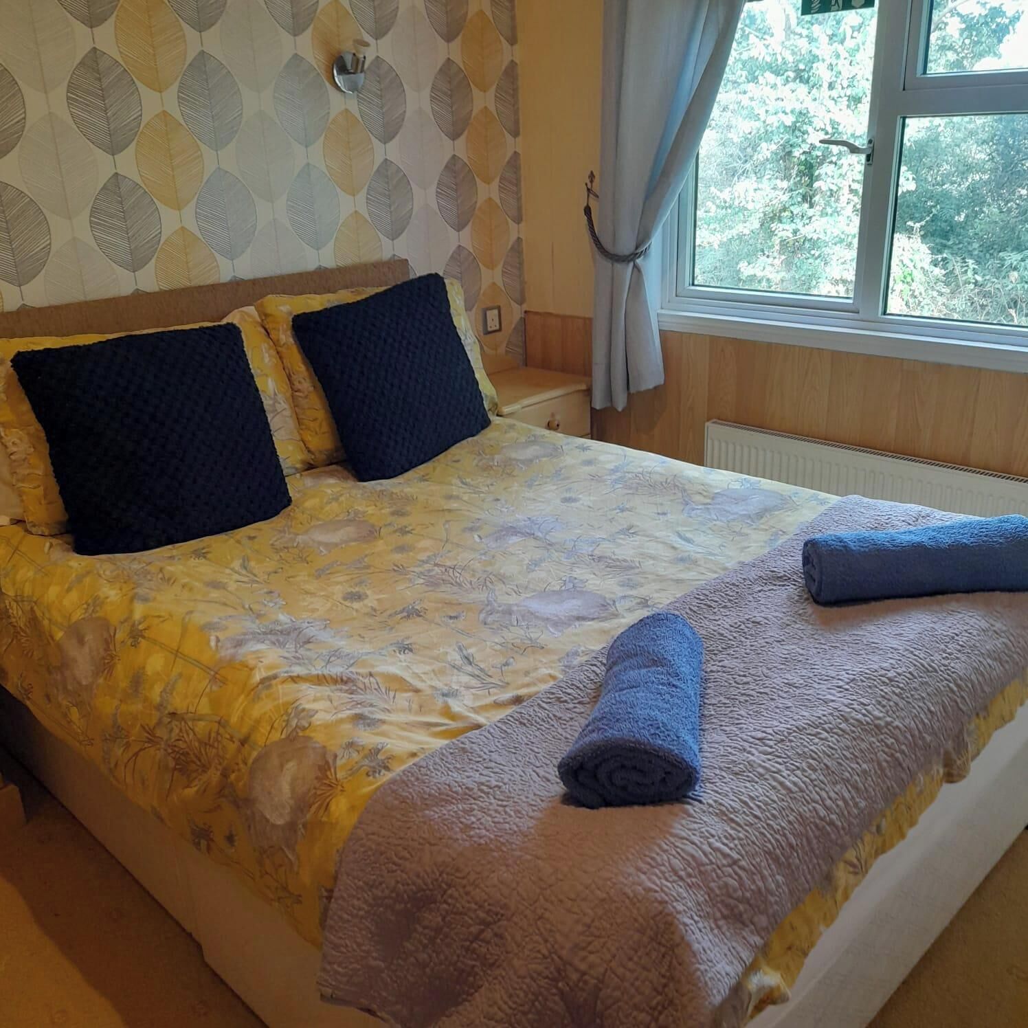 A bed with a floral bedspread, two dark pillows, and a rolled towel.
