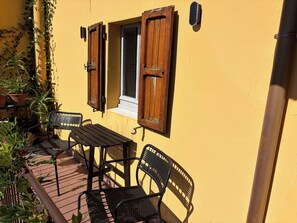 Ein Sitzbereich im Freien mit Tisch und Stühlen, ein Fenster mit Holzläden und Blumentöpfen.