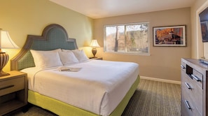 Una habitación de hotel con una cama, mesitas de noche, un aparador, un ventanal con persianas y un cuadro en la pared.