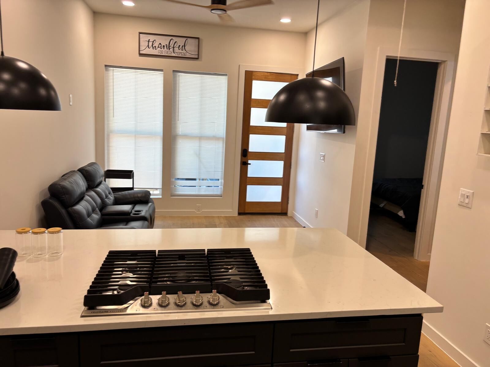 A modern kitchen with a central island, black pendant lights, and a black leather sofa in the adjacent living area.