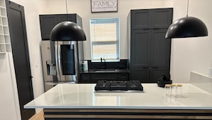 Una cocina moderna con una isla central, lámparas colgantes negras, un refrigerador de acero inoxidable y un ventanal con persianas.
