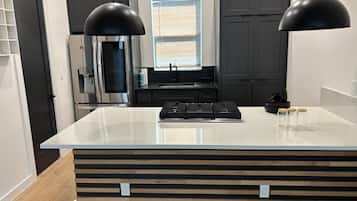 Una cocina moderna con una isla central, lámparas colgantes negras, un refrigerador de acero inoxidable y un ventanal con persianas.