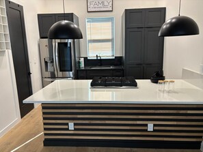 Een moderne keuken met een centraal eiland, zwarte hanglampen, een roestvrijstalen koelkast en een raam met jaloezieën.