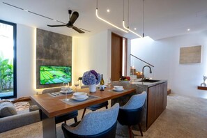 A modern dining area with a wooden table, two chairs, a TV mounted on the wall, and a kitchen in the background. - Pantai Lima 2BR Villa - Rooftop - 500m to Beach (Badung)