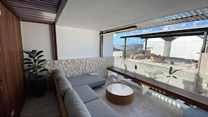 A modern living room with a stone accent wall, a grey sectional sofa, a round coffee table, and a view of construction work outside. - Pantai Lima 2BR Villa - Rooftop - 500m to Beach (Badung)