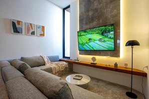 A modern living room with a large flat-screen TV mounted on the wall, a grey sectional sofa, a small round coffee table, and a floor lamp. - Pantai Lima 2BR Villa - Rooftop - 500m to Beach (Badung)